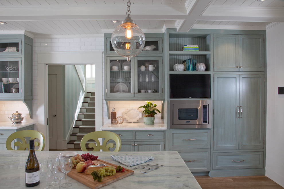 Diseño de cocina marinera, abierta, con armarios estilo shaker, puertas de armario azules, encimera de mármol, salpicadero blanco, salpicadero de azulejos tipo metro, electrodomésticos de acero inoxidable, suelo de madera en tonos medios y una isla