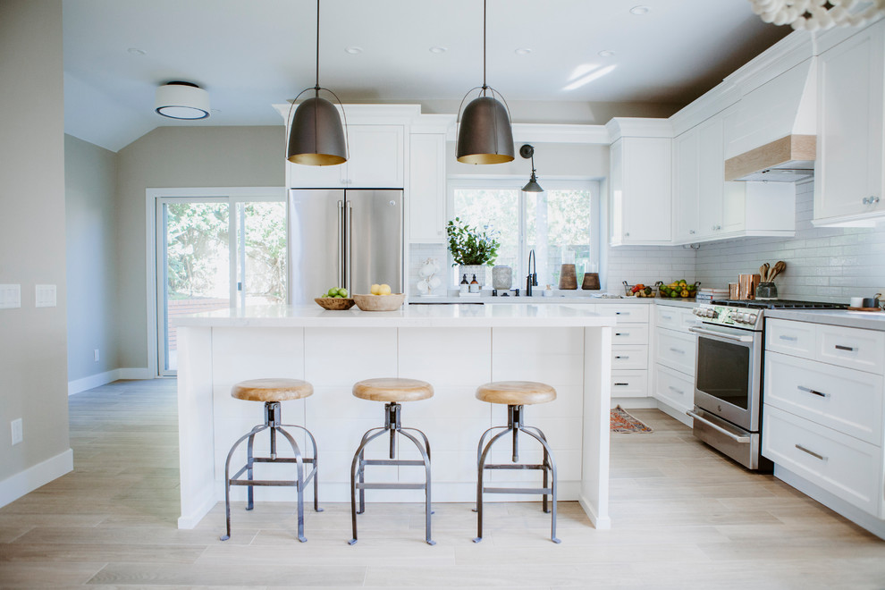 Photo of a medium sized beach style l-shaped open plan kitchen in San Diego with a submerged sink, shaker cabinets, white cabinets, engineered stone countertops, white splashback, metro tiled splashback, stainless steel appliances, ceramic flooring, an island and brown floors.