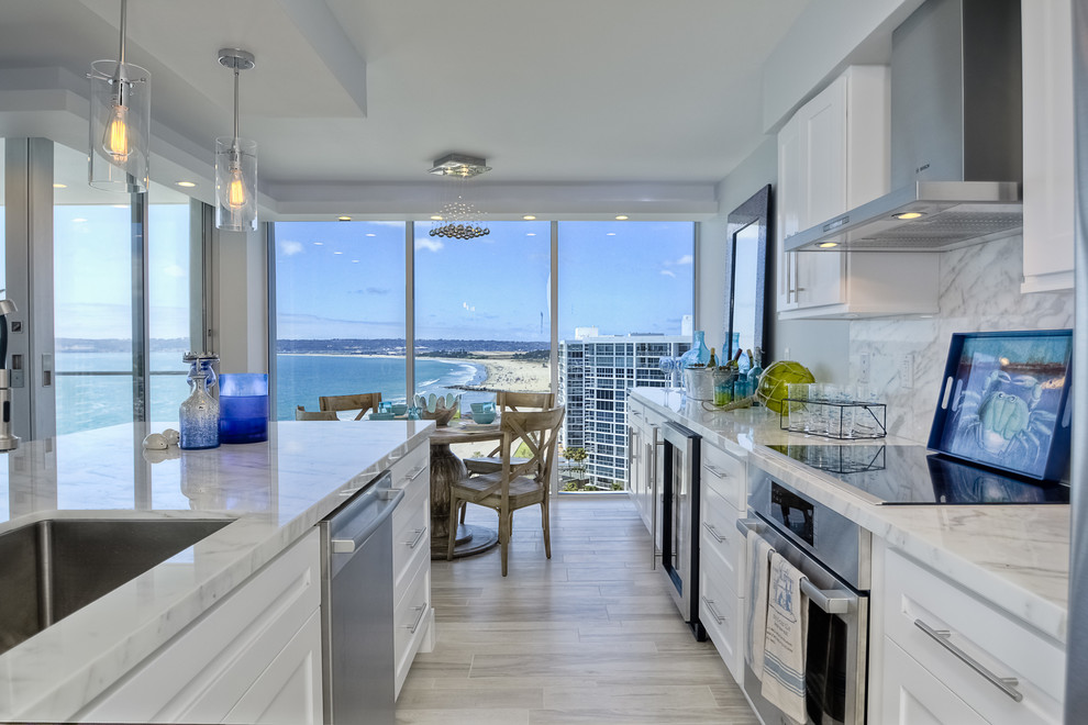 Small coastal galley open plan kitchen in San Diego with a single-bowl sink, shaker cabinets, white cabinets, marble worktops, stone slab splashback, stainless steel appliances, light hardwood flooring and an island.