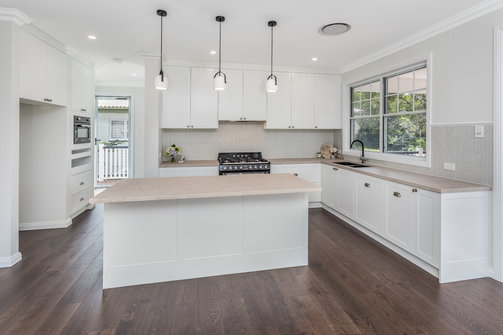 Photo of a medium sized classic kitchen pantry in Brisbane with a double-bowl sink, shaker cabinets, white cabinets, engineered stone countertops, beige splashback, ceramic splashback, black appliances, an island, brown floors and pink worktops.