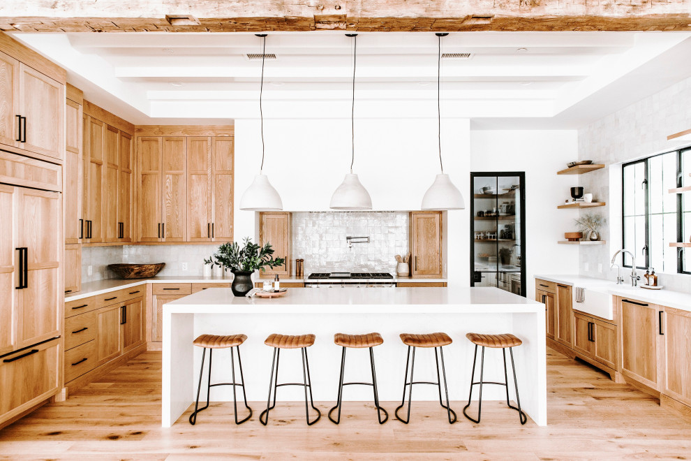 Photo of a country u-shaped kitchen in Phoenix with a belfast sink, shaker cabinets, medium wood cabinets, white splashback, stainless steel appliances, medium hardwood flooring, an island, brown floors and white worktops.