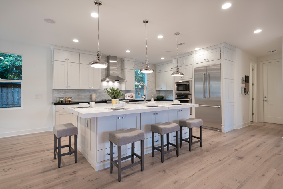 Photo of a large rural l-shaped kitchen/diner in Orange County with a belfast sink, shaker cabinets, white cabinets, engineered stone countertops, grey splashback, marble splashback, stainless steel appliances, light hardwood flooring and an island.