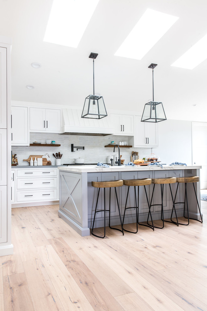 Inspiration for a large cottage light wood floor open concept kitchen remodel in Los Angeles with a farmhouse sink, white cabinets, marble countertops, gray backsplash, black appliances and an island
