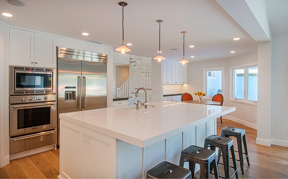 Inspiration for a large coastal l-shaped light wood floor open concept kitchen remodel in Orange County with a farmhouse sink, shaker cabinets, white cabinets, quartz countertops, white backsplash, mosaic tile backsplash, stainless steel appliances and an island