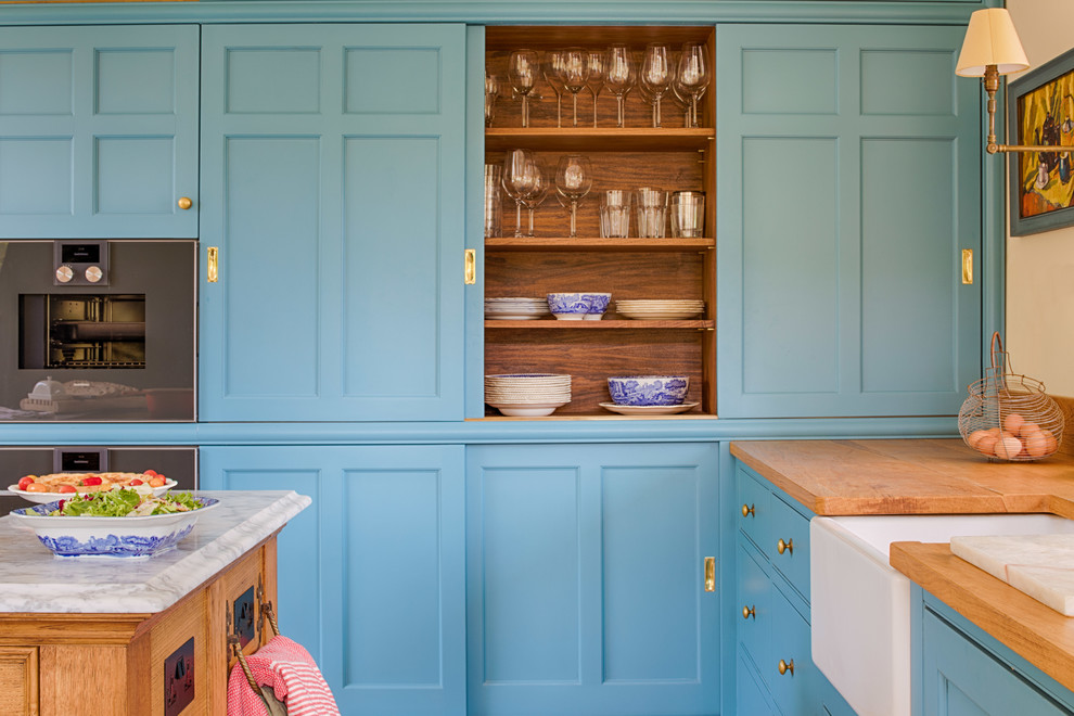 Wohnküche in L-Form mit Landhausspüle, blauen Schränken, Arbeitsplatte aus Holz, Elektrogeräten mit Frontblende, Terrakottaboden, Kücheninsel, brauner Arbeitsplatte und Schrankfronten im Shaker-Stil in Gloucestershire