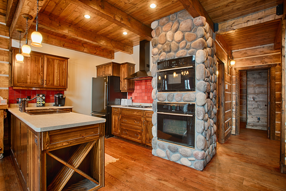 Exemple d'une cuisine montagne en bois brun avec un évier 2 bacs, un placard à porte shaker, une crédence rouge, un électroménager noir, un sol en bois brun, une péninsule, un sol marron et un plan de travail gris.
