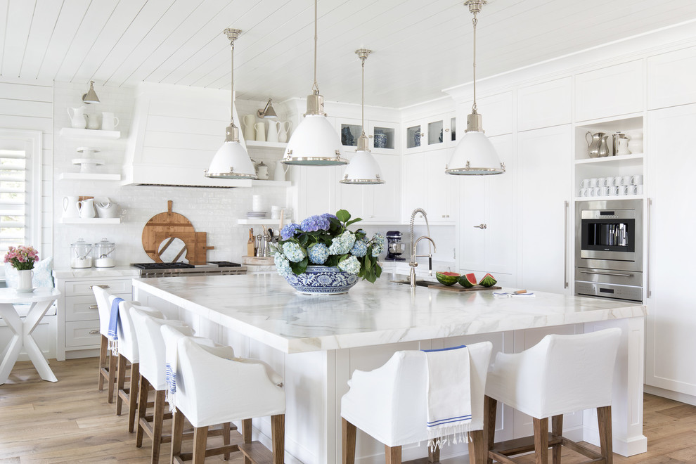 Maritim inredning av ett vit vitt l-kök, med marmorbänkskiva, en köksö, en undermonterad diskho, luckor med infälld panel, vita skåp, vitt stänkskydd, rostfria vitvaror, mellanmörkt trägolv och brunt golv
