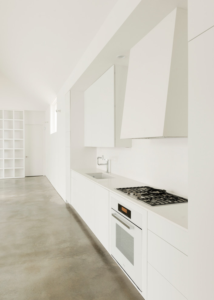 Example of a mid-sized trendy single-wall concrete floor open concept kitchen design in Houston with an undermount sink, flat-panel cabinets, white cabinets, white appliances, solid surface countertops, no island and white backsplash