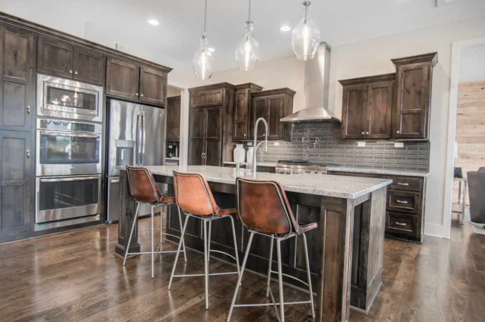 Example of a large urban single-wall dark wood floor open concept kitchen design in Kansas City with a farmhouse sink, recessed-panel cabinets, dark wood cabinets, granite countertops, gray backsplash, ceramic backsplash, stainless steel appliances and an island