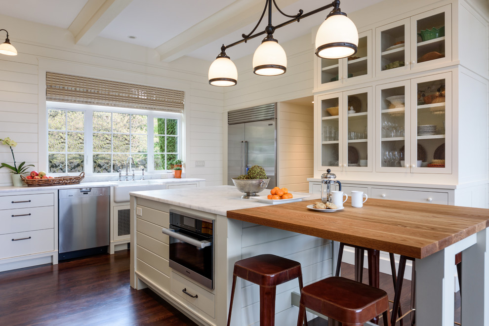 Lantlig inredning av ett vit vitt kök, med en rustik diskho, luckor med glaspanel, vita skåp, fönster som stänkskydd, rostfria vitvaror, mörkt trägolv och brunt golv