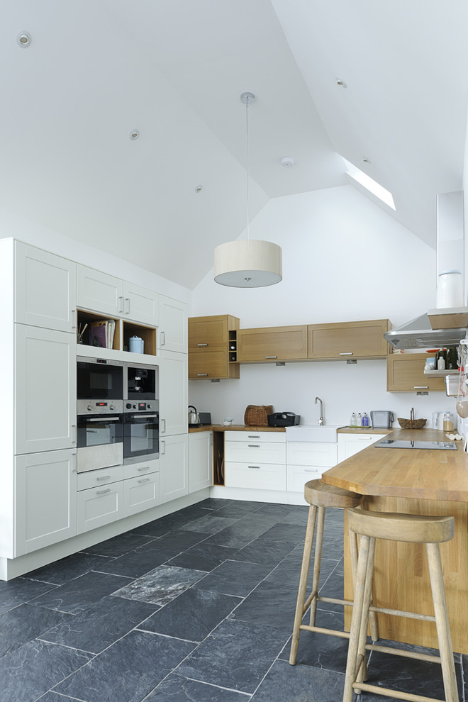 Photo of a medium sized country u-shaped kitchen in Other with a belfast sink, recessed-panel cabinets, white cabinets, wood worktops, stainless steel appliances, a breakfast bar, black floors and beige worktops.