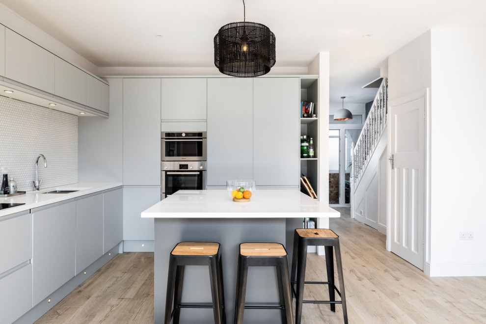 Contemporary kitchen in Hertfordshire with a submerged sink, flat-panel cabinets, grey cabinets, white splashback, stainless steel appliances, light hardwood flooring, an island and white worktops.