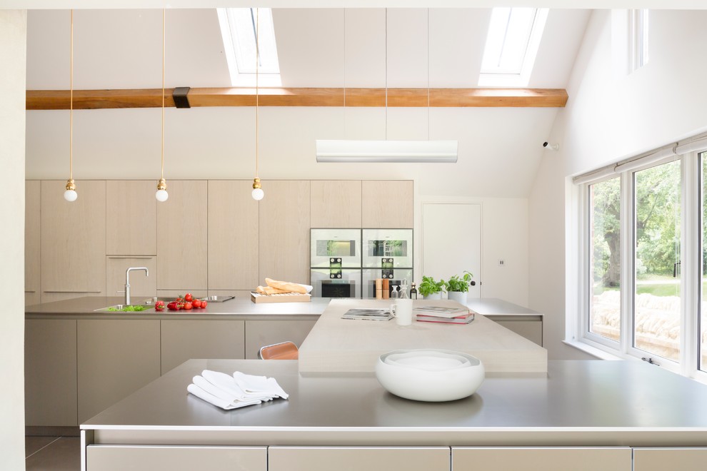 Moderne Küche mit flächenbündigen Schrankfronten, hellen Holzschränken, zwei Kücheninseln, Edelstahl-Arbeitsplatte und Küchengeräten aus Edelstahl in Oxfordshire