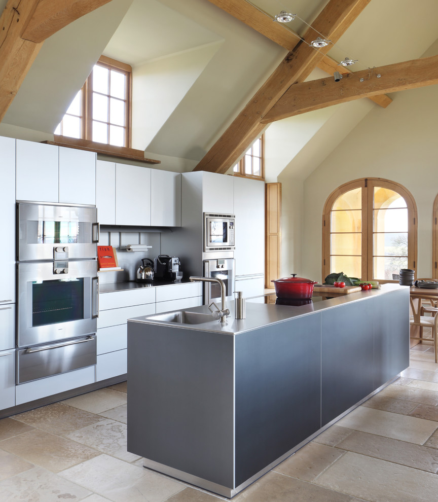Photo of a rural kitchen/diner in London with an integrated sink, flat-panel cabinets, white cabinets, stainless steel worktops, metallic splashback, stainless steel appliances, an island and beige floors.