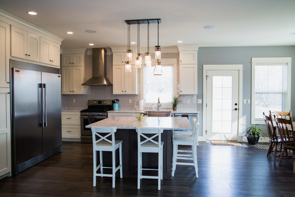Example of an arts and crafts l-shaped laminate floor and brown floor eat-in kitchen design in Grand Rapids with a farmhouse sink, shaker cabinets, white cabinets, quartz countertops, gray backsplash, ceramic backsplash, stainless steel appliances, an island and white countertops