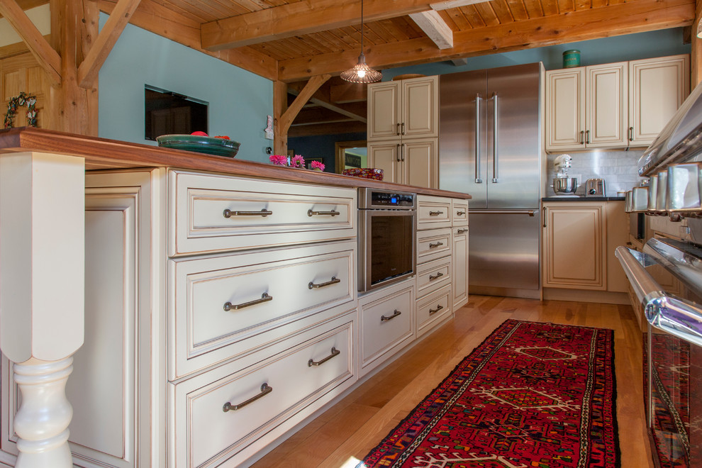 Lantlig inredning av ett mellanstort kök, med en rustik diskho, luckor med upphöjd panel, gula skåp, bänkskiva i täljsten, grått stänkskydd, stänkskydd i stenkakel, rostfria vitvaror, mellanmörkt trägolv och en köksö