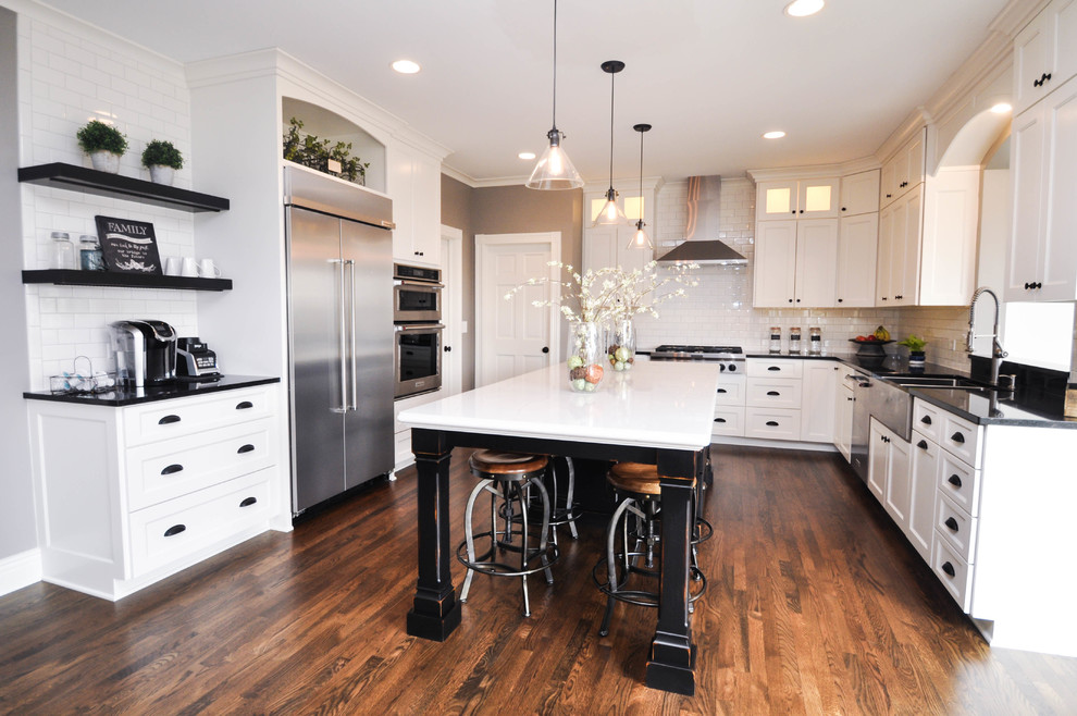 This is an example of a country u-shaped kitchen in Minneapolis with shaker cabinets, white cabinets, engineered stone countertops, metro tiled splashback, stainless steel appliances, an island, a belfast sink, medium hardwood flooring and brown floors.