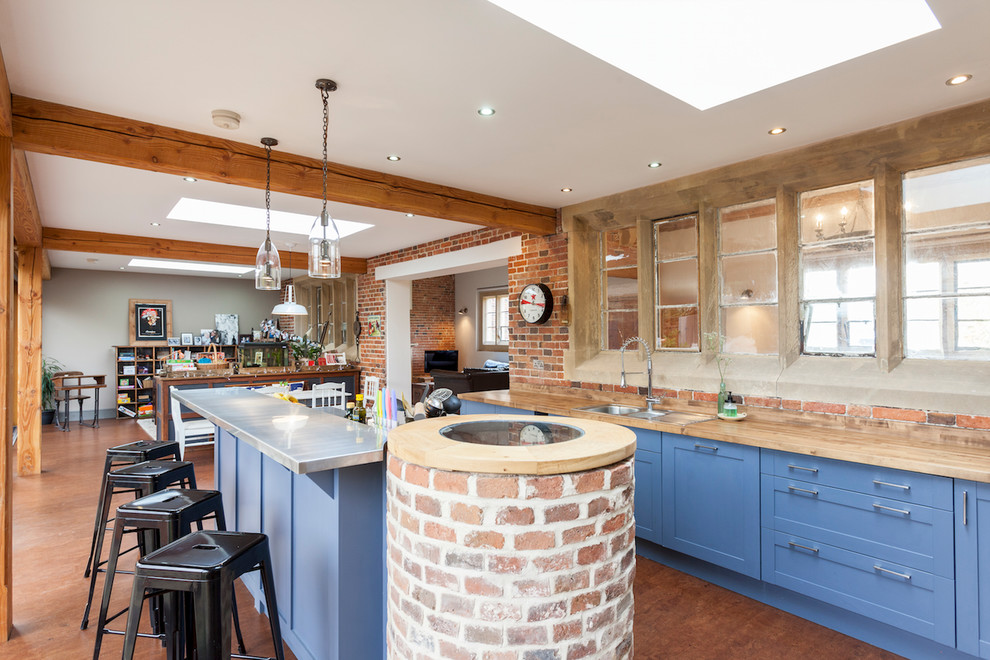 Inspiration for a farmhouse galley open plan kitchen in London with a double-bowl sink, shaker cabinets, blue cabinets, wood worktops, red splashback, brick splashback, an island and brown floors.