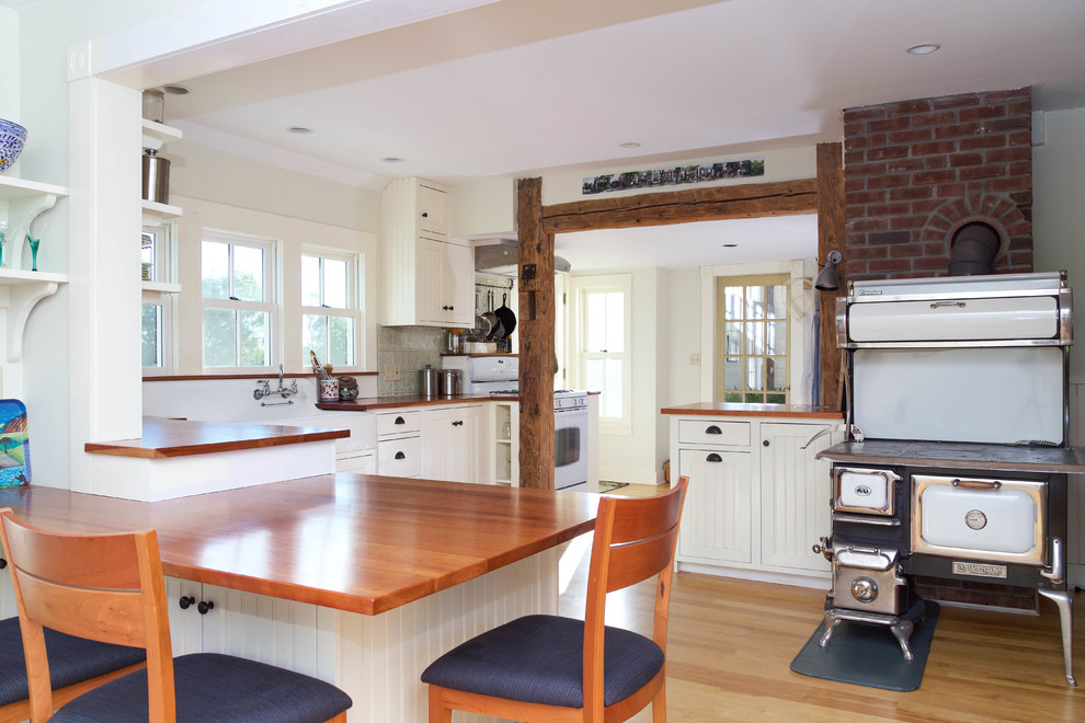 Foto på ett lantligt brun kök, med en rustik diskho, skåp i shakerstil, vita skåp, träbänkskiva, grått stänkskydd, svarta vitvaror, ljust trägolv, en halv köksö och beiget golv