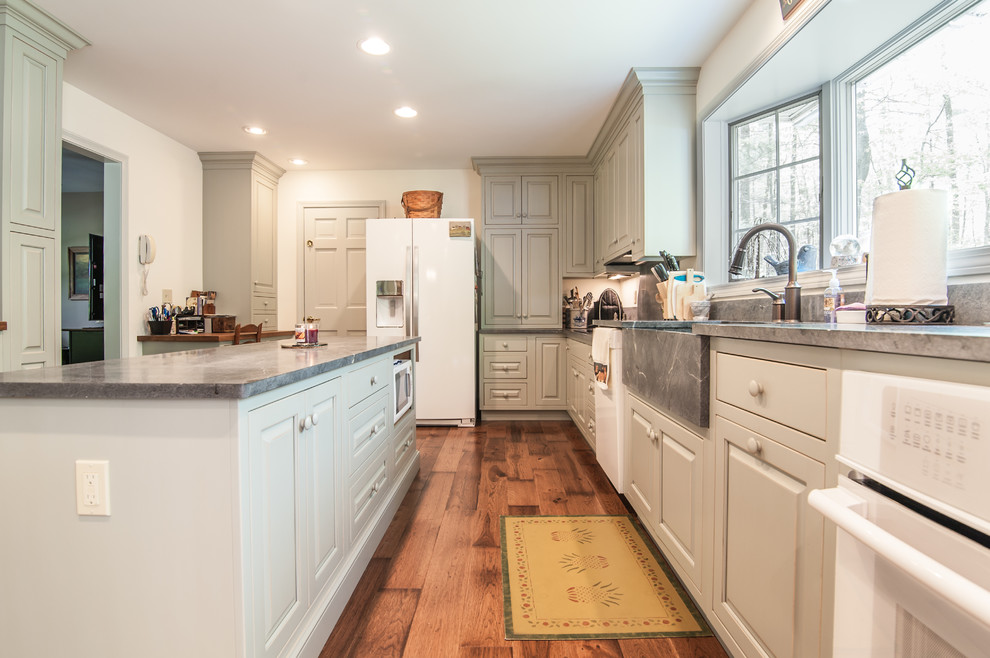 Photo of a large country l-shaped kitchen/diner in Providence with a belfast sink, raised-panel cabinets, green cabinets, soapstone worktops, white appliances, medium hardwood flooring and an island.