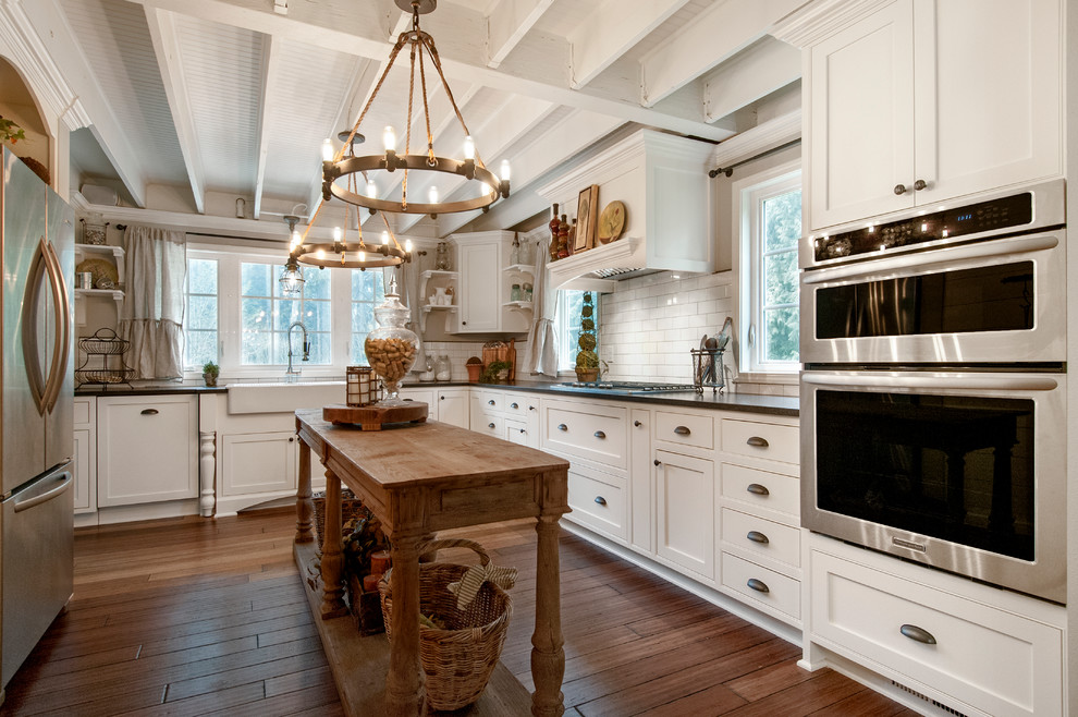 Photo of a rural u-shaped kitchen/diner in Seattle with a belfast sink, shaker cabinets, white cabinets, white splashback, metro tiled splashback, stainless steel appliances, engineered stone countertops, medium hardwood flooring, an island and brown floors.