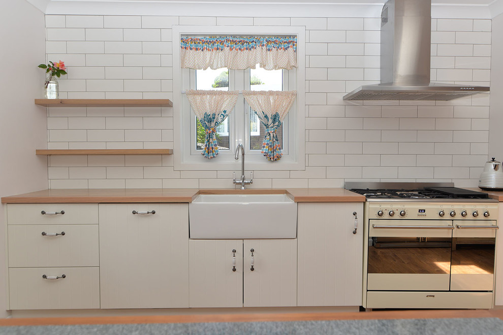 Photo of a small rural single-wall kitchen/diner in Auckland with a belfast sink, shaker cabinets, white cabinets, wood worktops, white splashback, ceramic splashback, coloured appliances, medium hardwood flooring, no island and brown floors.