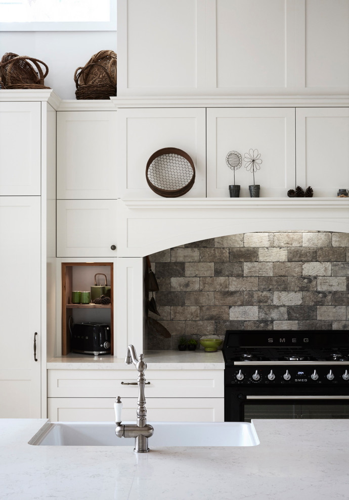Photo of a large rural galley kitchen pantry in Sydney with a belfast sink, shaker cabinets, white cabinets, engineered stone countertops, grey splashback, stone tiled splashback, black appliances, travertine flooring, an island, grey floors and white worktops.