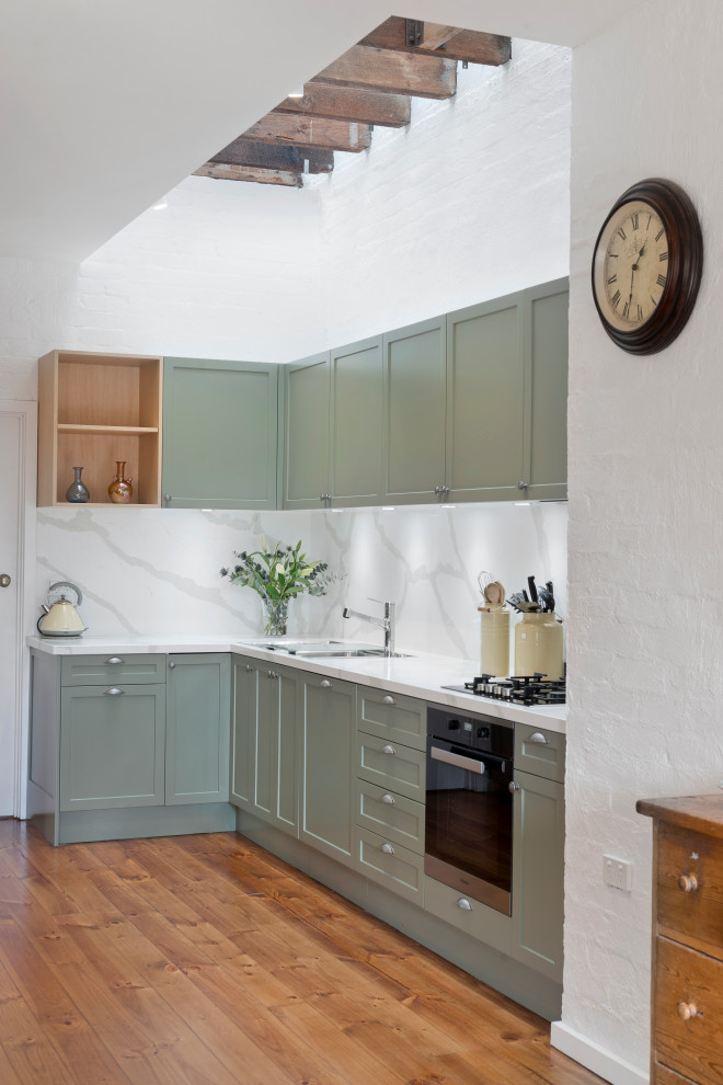 Example of a mid-sized transitional l-shaped medium tone wood floor and brown floor eat-in kitchen design in Melbourne with a drop-in sink, shaker cabinets, green cabinets, quartz countertops, white backsplash, stone slab backsplash, black appliances, an island and white countertops