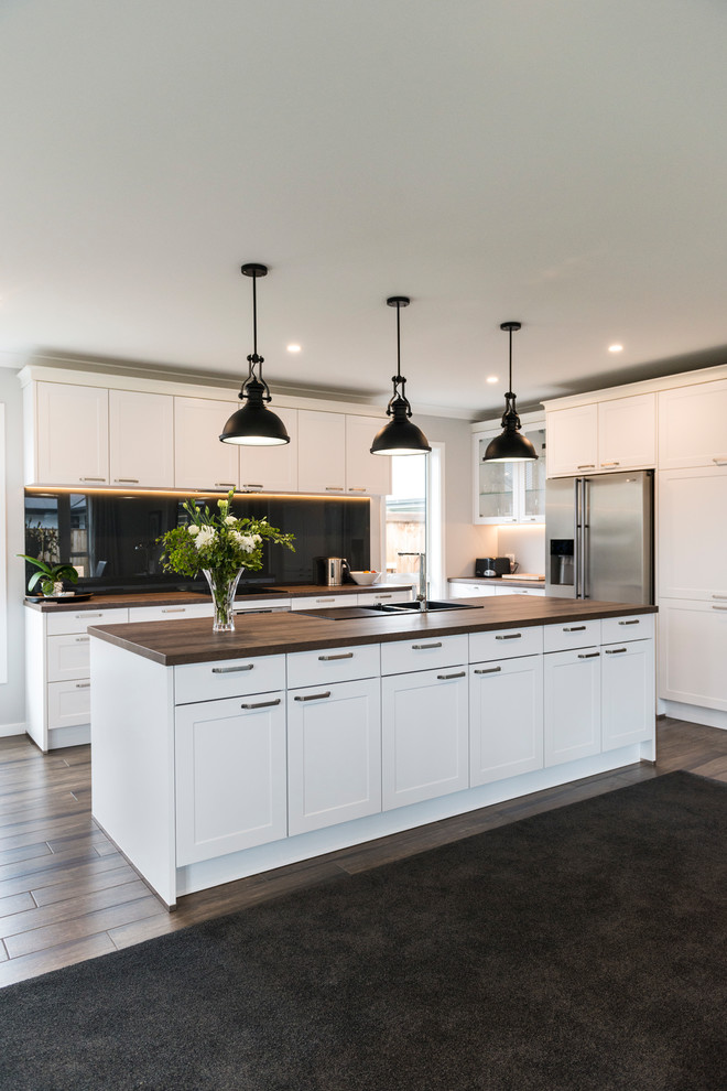 This is an example of a large traditional l-shaped kitchen pantry in Christchurch with a belfast sink, shaker cabinets, white cabinets, laminate countertops, black splashback, glass sheet splashback, stainless steel appliances, vinyl flooring, an island and brown floors.