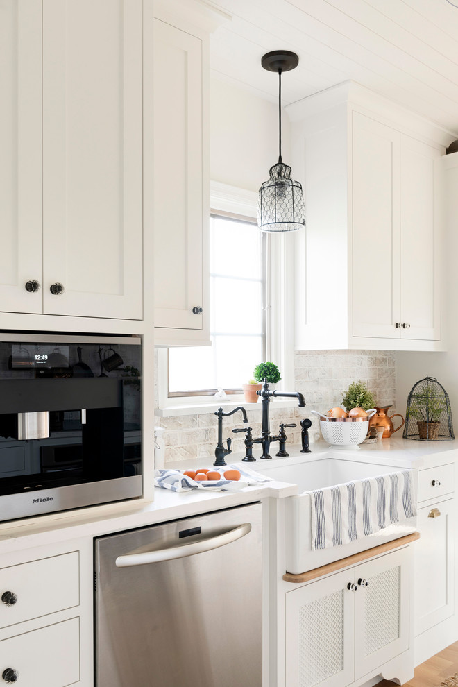 Example of a farmhouse u-shaped light wood floor and beige floor eat-in kitchen design in Minneapolis with a farmhouse sink, white cabinets, white backsplash, brick backsplash, stainless steel appliances, an island and white countertops