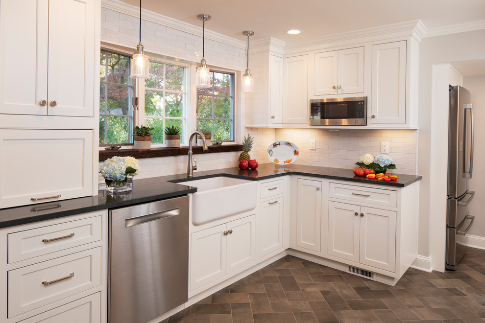Example of a mid-sized transitional l-shaped ceramic tile and brown floor enclosed kitchen design in New York with a farmhouse sink, shaker cabinets, white cabinets, quartz countertops, white backsplash, subway tile backsplash, stainless steel appliances, no island and black countertops