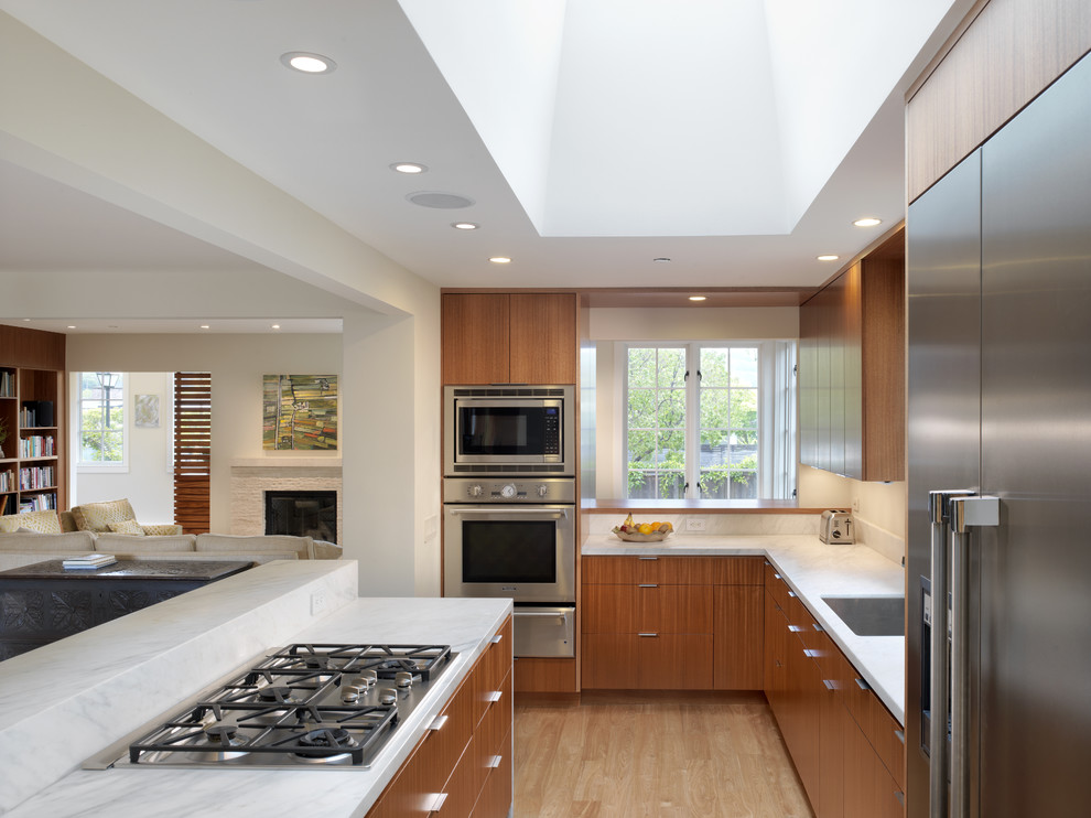На фото: п-образная кухня-гостиная в стиле модернизм с техникой из нержавеющей стали, врезной мойкой, плоскими фасадами и фасадами цвета дерева среднего тона