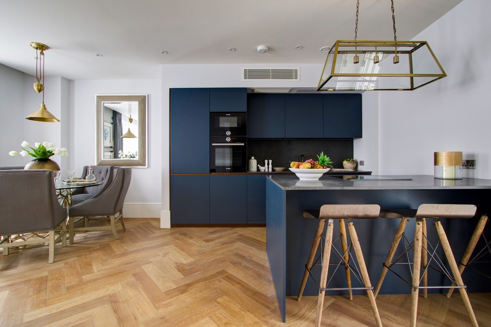 Moderne Wohnküche mit flächenbündigen Schrankfronten, blauen Schränken, Küchenrückwand in Schwarz, schwarzen Elektrogeräten, hellem Holzboden und Halbinsel in London