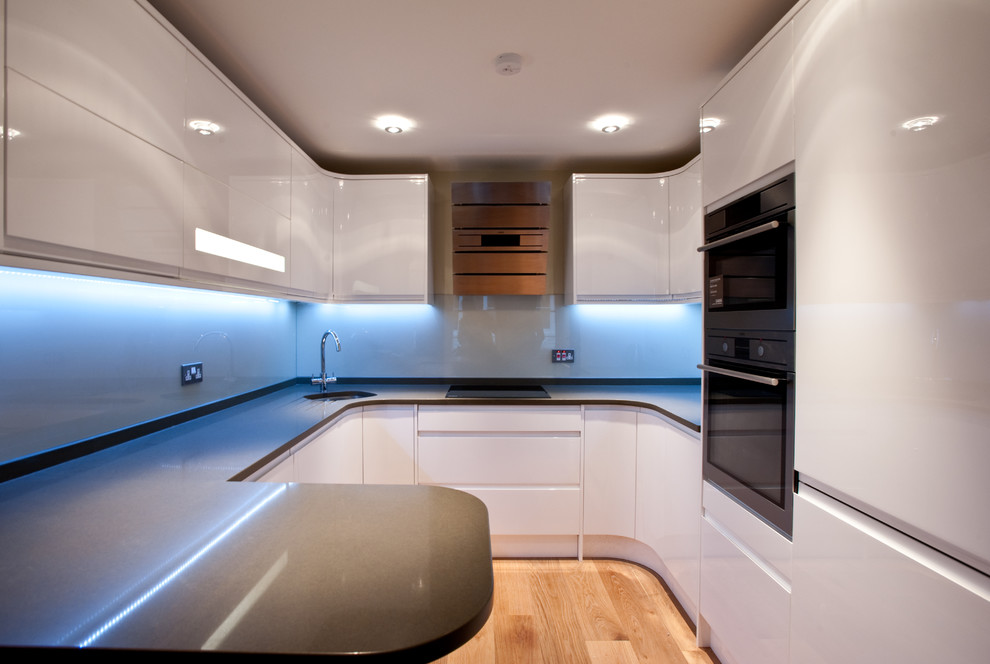 Photo of a small modern galley enclosed kitchen in London with a submerged sink, flat-panel cabinets, white cabinets, granite worktops, white splashback, a breakfast bar, stone slab splashback, stainless steel appliances and light hardwood flooring.