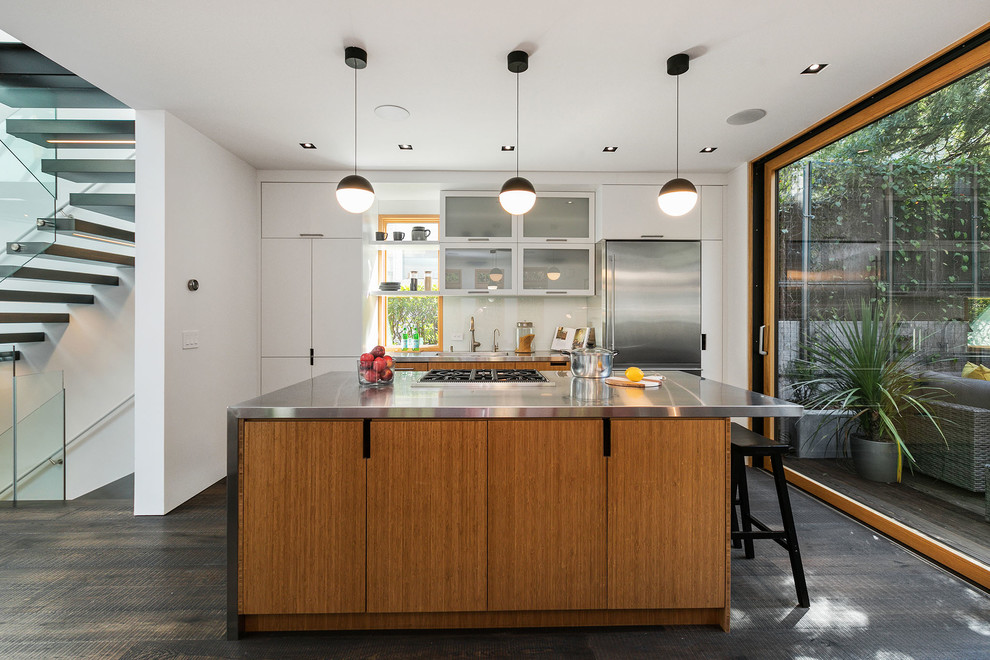 Inredning av ett eklektiskt grå linjärt grått kök med öppen planlösning, med släta luckor, skåp i mellenmörkt trä, bänkskiva i rostfritt stål, vitt stänkskydd, stänkskydd i sten, rostfria vitvaror, mörkt trägolv, en köksö, brunt golv och en integrerad diskho