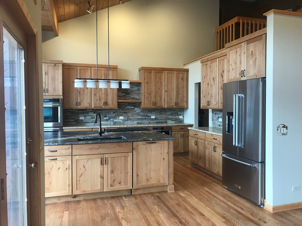 Photo of a large rustic l-shaped open plan kitchen in Chicago with a submerged sink, shaker cabinets, medium wood cabinets, granite worktops, green splashback, slate splashback, stainless steel appliances, medium hardwood flooring, an island, brown floors and black worktops.