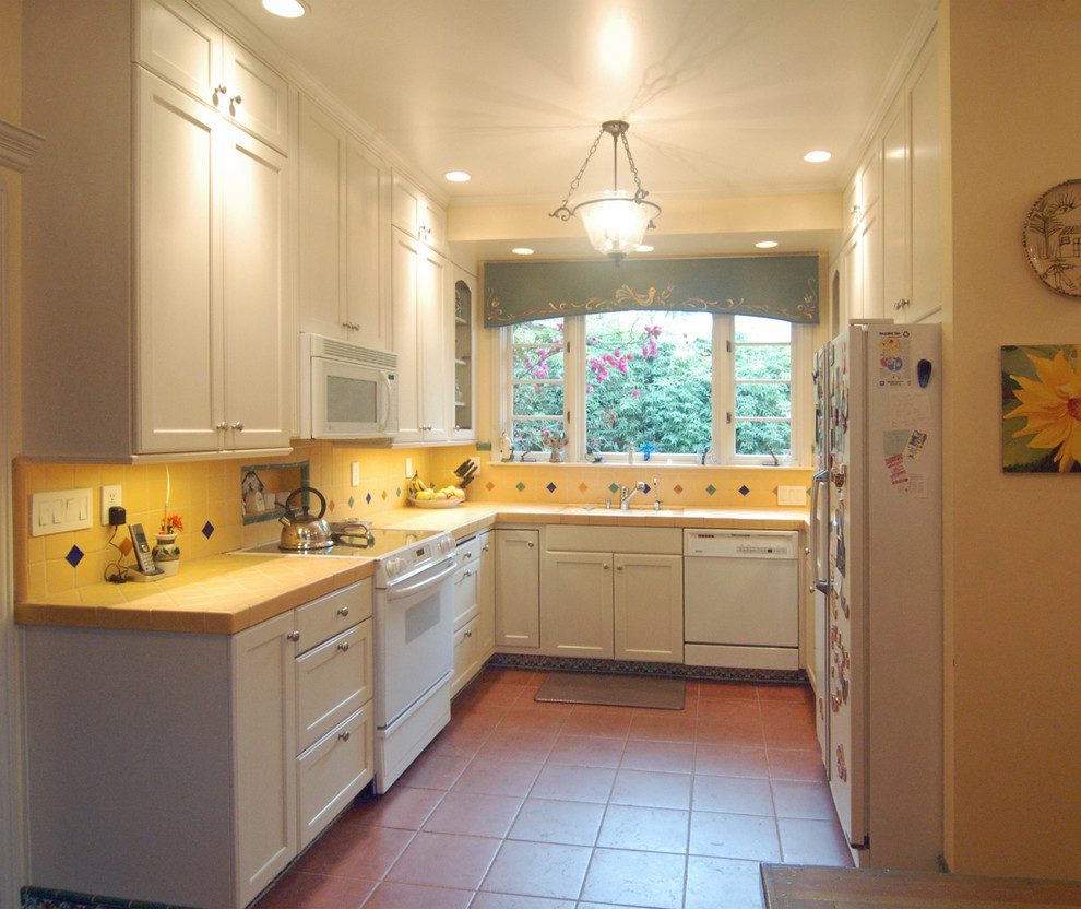 Exempel på ett litet amerikanskt kök, med en undermonterad diskho, luckor med infälld panel, vita skåp, kaklad bänkskiva, gult stänkskydd, stänkskydd i keramik, vita vitvaror och klinkergolv i keramik