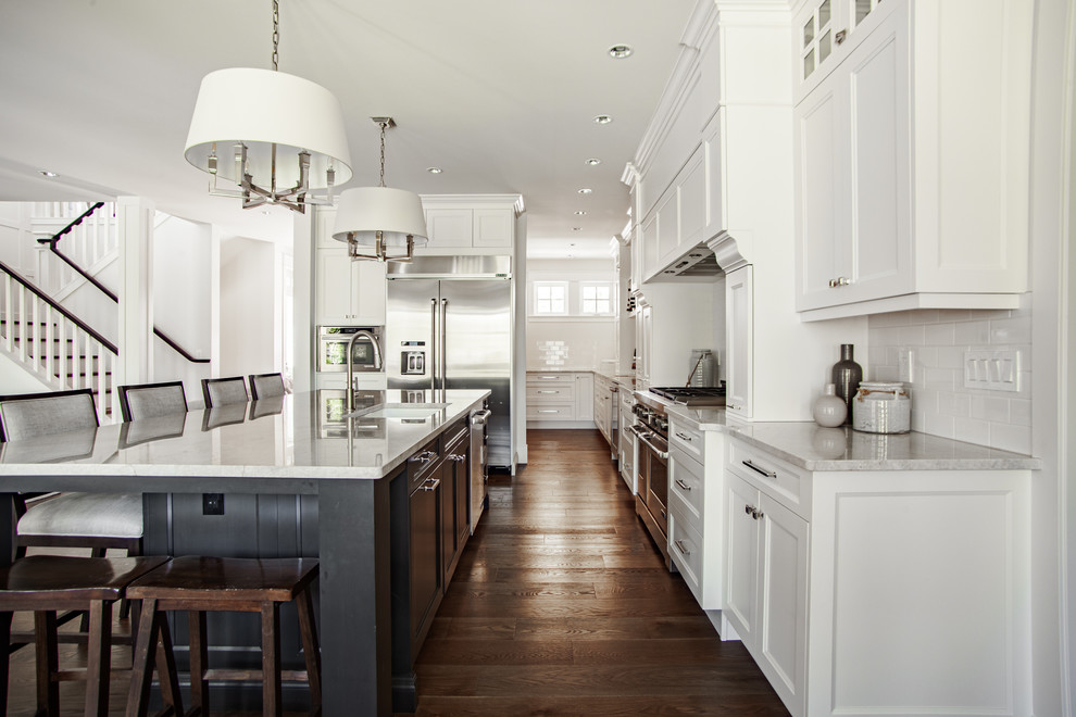 This is an example of a large traditional l-shaped open plan kitchen in Vancouver with a submerged sink, shaker cabinets, white cabinets, quartz worktops, white splashback, ceramic splashback, stainless steel appliances, dark hardwood flooring, an island, brown floors and grey worktops.