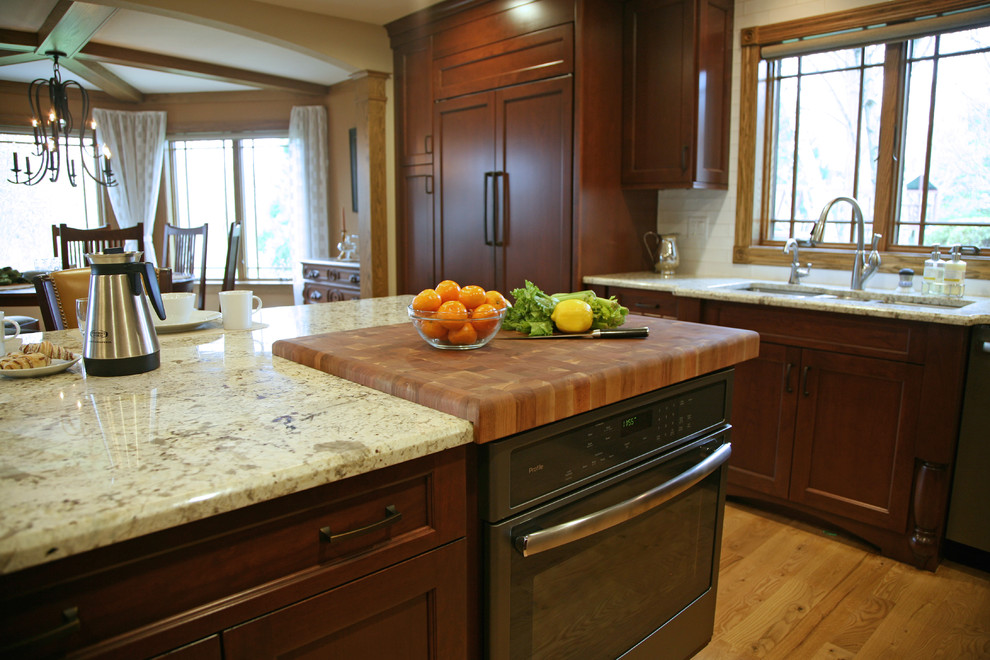 Exempel på ett stort amerikanskt kök, med en trippel diskho, släta luckor, skåp i mellenmörkt trä, granitbänkskiva, vitt stänkskydd, stänkskydd i keramik, rostfria vitvaror, ljust trägolv och en köksö