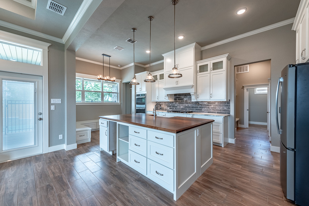Eat-in kitchen - large craftsman ceramic tile and brown floor eat-in kitchen idea in Other with a farmhouse sink, shaker cabinets, white cabinets, wood countertops, gray backsplash, brick backsplash, stainless steel appliances, an island and beige countertops