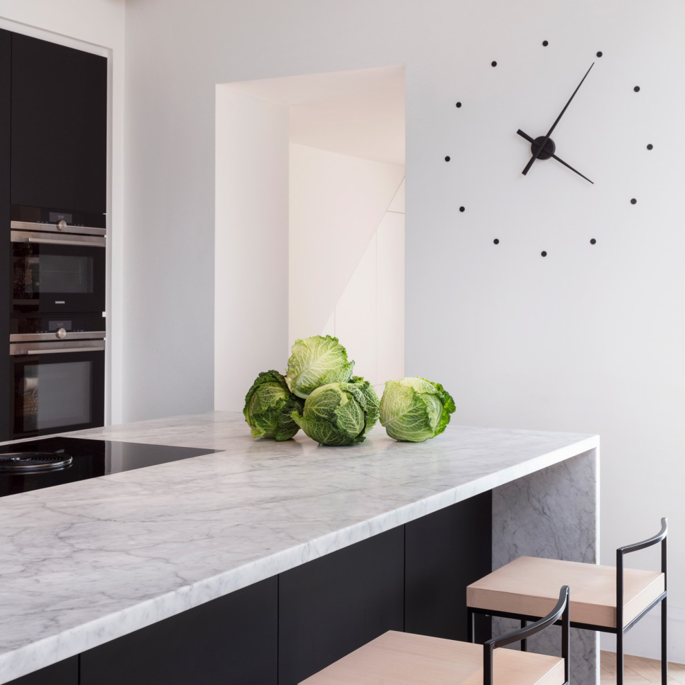 Minimalistisk inredning av ett mellanstort grå linjärt grått kök och matrum, med en undermonterad diskho, släta luckor, svarta skåp, marmorbänkskiva, grått stänkskydd, stänkskydd i marmor, svarta vitvaror, ljust trägolv, en köksö och brunt golv