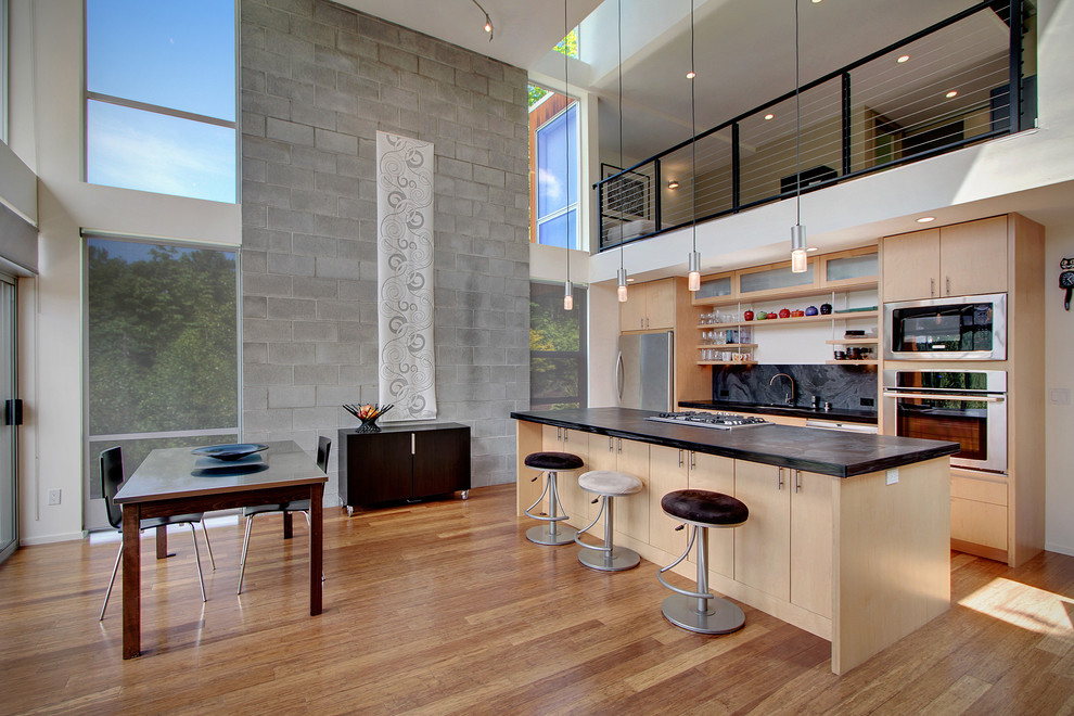 Источник вдохновения для домашнего уюта: параллельная кухня в стиле модернизм с черным фартуком, фартуком из каменной плиты, обеденным столом, плоскими фасадами, светлыми деревянными фасадами и техникой из нержавеющей стали