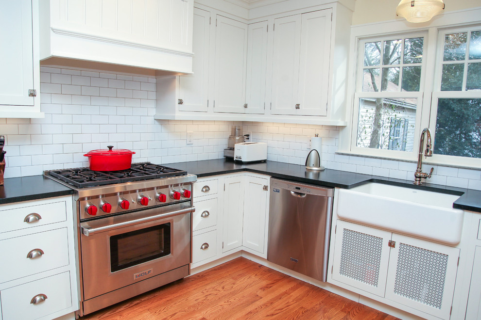 Example of a mid-sized country l-shaped light wood floor enclosed kitchen design in New York with a farmhouse sink, shaker cabinets, white cabinets, granite countertops, white backsplash, subway tile backsplash, stainless steel appliances and no island