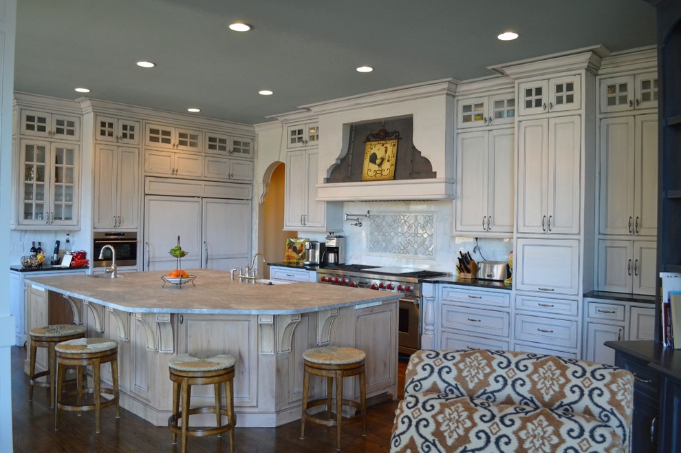 Example of a large transitional l-shaped medium tone wood floor eat-in kitchen design in Atlanta with a farmhouse sink, shaker cabinets, distressed cabinets, solid surface countertops, white backsplash, stone tile backsplash, stainless steel appliances and an island