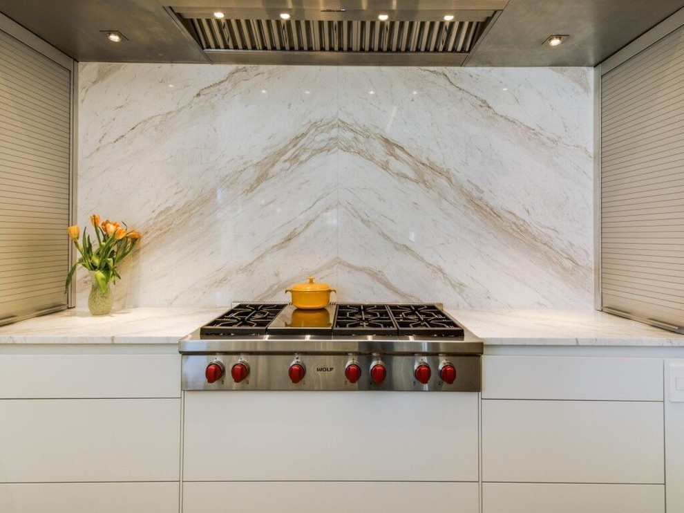Example of a small trendy l-shaped limestone floor enclosed kitchen design in Dallas with an undermount sink, flat-panel cabinets, white cabinets, marble countertops, white backsplash, glass sheet backsplash, stainless steel appliances and an island