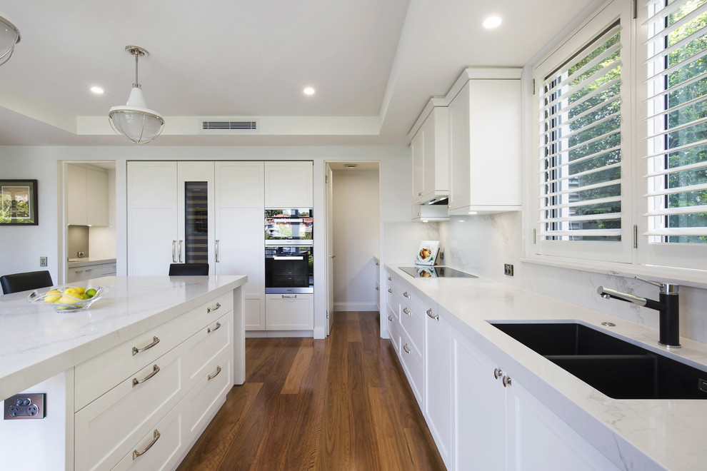 This is an example of a large classic u-shaped open plan kitchen in Sydney with a submerged sink, shaker cabinets, white cabinets, engineered stone countertops, white splashback, stone slab splashback, black appliances, medium hardwood flooring and an island.