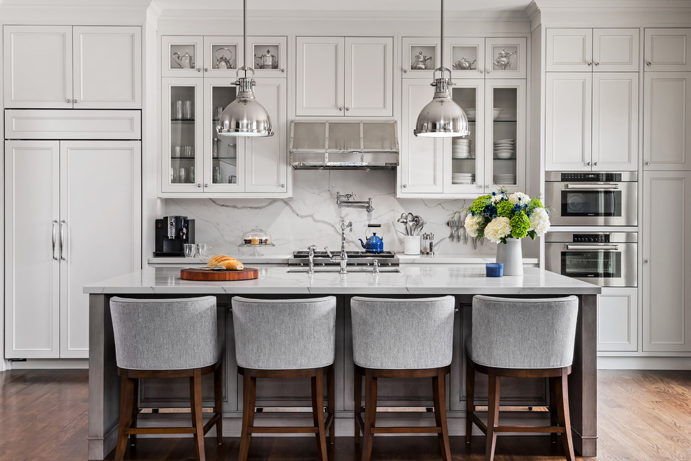 This is an example of a large traditional grey and white galley kitchen in Toronto with a submerged sink, recessed-panel cabinets, white cabinets, white splashback, integrated appliances, medium hardwood flooring, an island and brown floors.