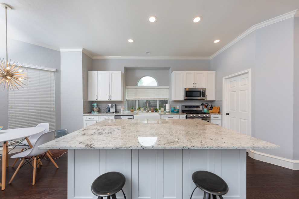 Example of a mid-sized transitional single-wall medium tone wood floor and brown floor open concept kitchen design in Dallas with a farmhouse sink, shaker cabinets, white cabinets, granite countertops, gray backsplash, subway tile backsplash, stainless steel appliances, an island and white countertops