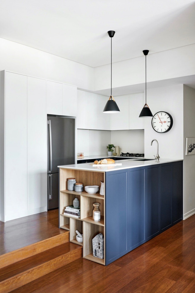 Foto på ett mellanstort funkis vit kök, med en undermonterad diskho, skåp i shakerstil, blå skåp, bänkskiva i kvarts, vitt stänkskydd, rostfria vitvaror, mellanmörkt trägolv, en halv köksö och brunt golv