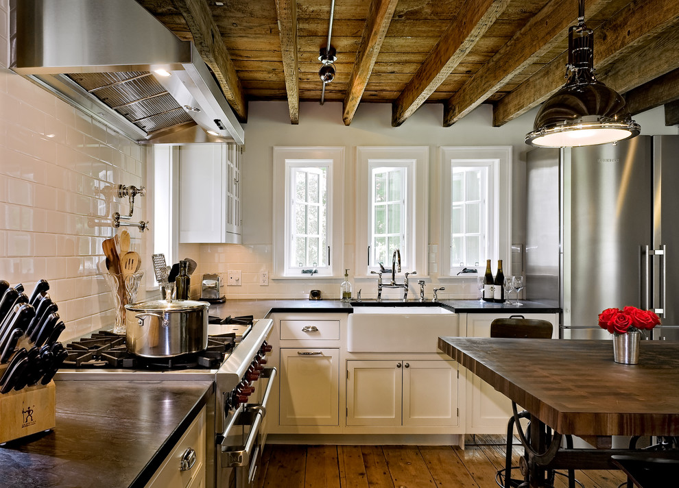 Bild på ett lantligt l-kök, med en rustik diskho, luckor med profilerade fronter, vita skåp, vitt stänkskydd, stänkskydd i tunnelbanekakel, rostfria vitvaror och bänkskiva i täljsten